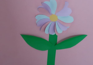 kolorowy kwiatek wykonany z niebieskich, różowych i fioletowych serduszek naklejonych jedne na drugich, dwoma zielonymi liściami i łodygą na różowej kartce